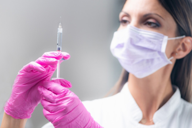A masked aesthetic practitioner in pink gloves carefully prepares a hyaluronic acid syringe, ready for a facial filler treatment.